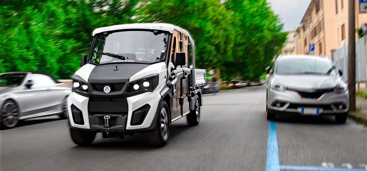 Alkè ATX Elektro-Nutzfahrzeug fährt durch eine Straße mit parkenden Autos und grünen Bäumen im Hintergrund.
