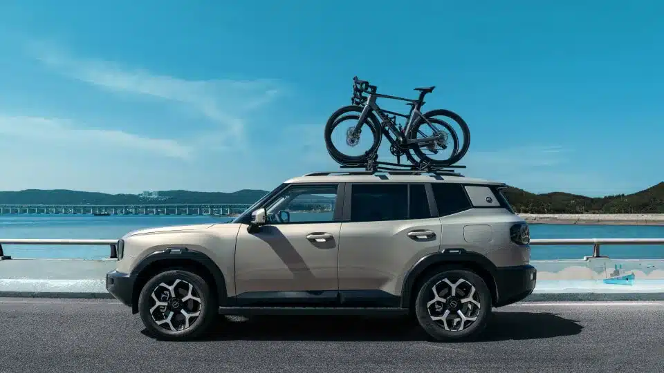 Beiger Jetour T1 i-DM SUV mit montiertem Fahrradträger auf dem Dach steht auf einer Küstenstraße vor blauem Himmel und Wasser.