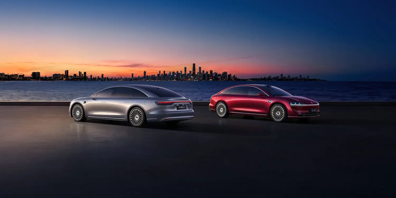 Zwei Xiangjie S9 Limousinen (silber und rot) stehen bei Sonnenuntergang vor einer Stadt am Wasser.