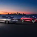 Zwei Xiangjie S9 Limousinen (silber und rot) stehen bei Sonnenuntergang vor einer Stadt am Wasser.