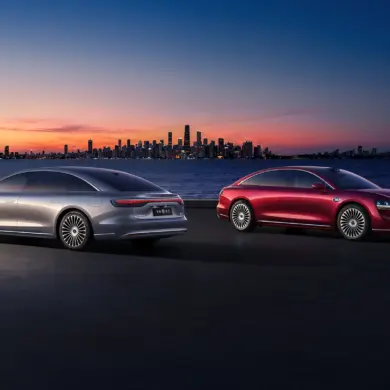 Zwei Xiangjie S9 Limousinen (silber und rot) stehen bei Sonnenuntergang vor einer Stadt am Wasser.