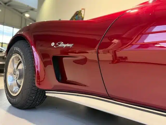 Nahaufnahme eines roten Corvette C3 Stingray Kotflügels mit Stingray-Emblem und Reifen in einer hellen Halle.