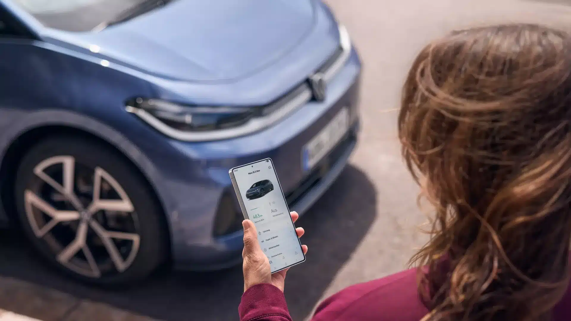 Person hält ein Smartphone mit Fahrzeug-App vor einem blauen Volkswagen ID.3 Neo.