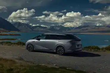 XPENG X9 fährt auf einer Uferstraße mit See und Bergen im Hintergrund.
