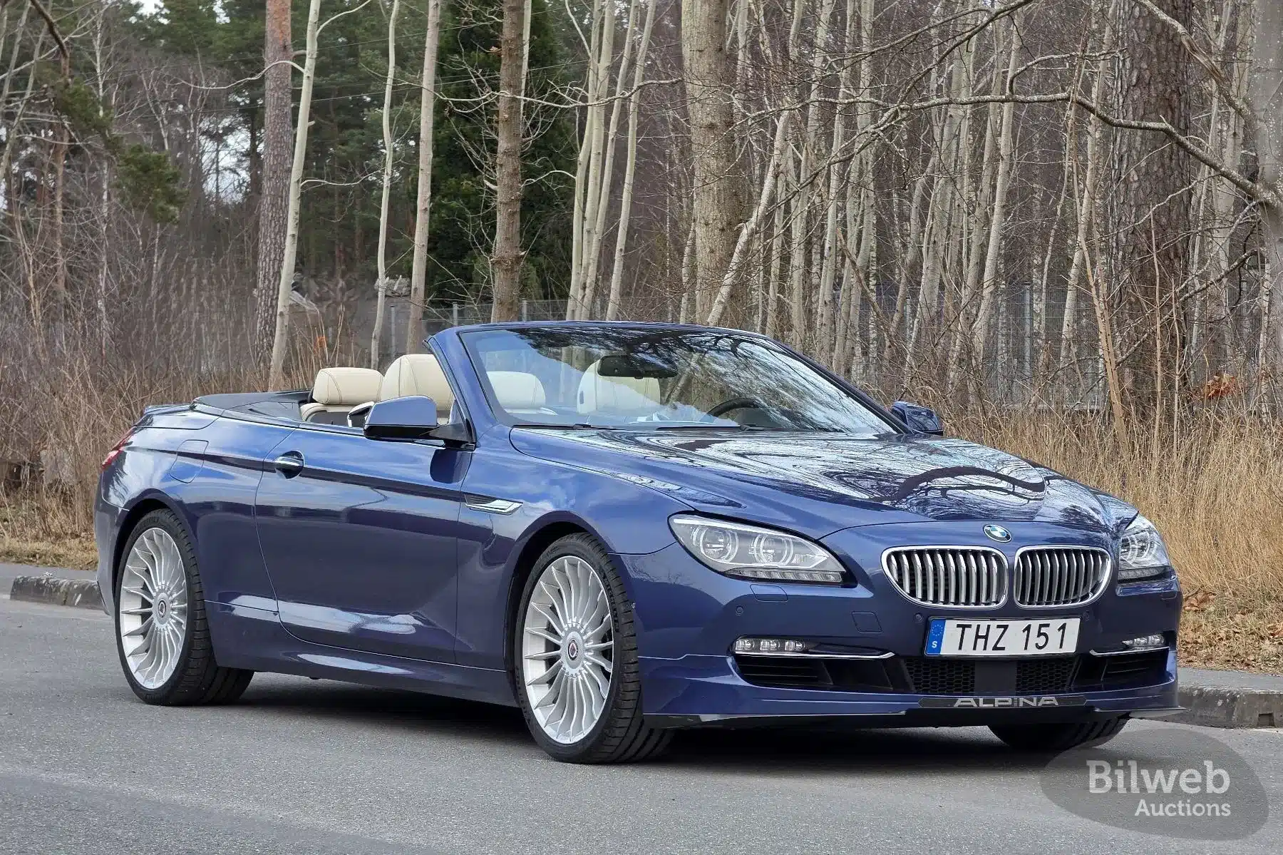 Blauer Alpina B6 Biturbo Cabriolet aus dem Jahr 2012 mit beigem Lederinterieur und geöffnetem Verdeck, seitlich fotografiert auf einer Straße vor bewaldetem Hintergrund.