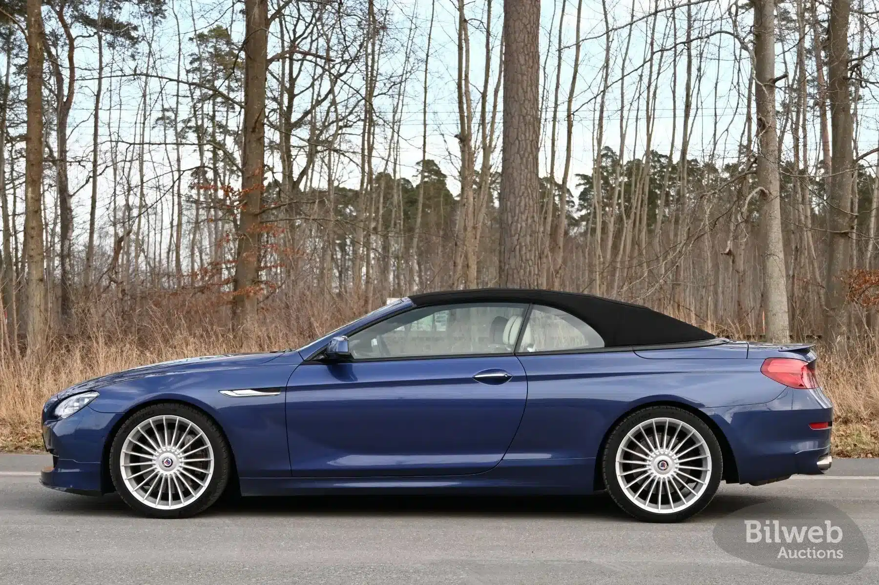 Blaues Alpina B6 Biturbo Cabriolet von 2012 mit geschlossenem Verdeck im Profil, geparkt am Waldrand.