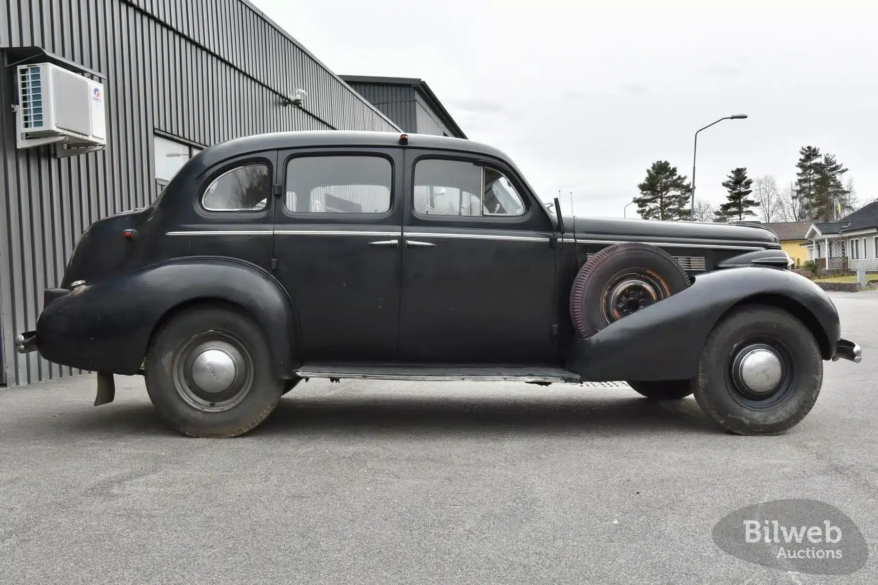 Schwarzer Buick Special Saloon Straight 8 Baujahr 1937 mit sichtbaren Gebrauchsspuren und fehlendem Ersatzrad an der Seite, aufgenommen auf einem Parkplatz vor einer Industriehalle.