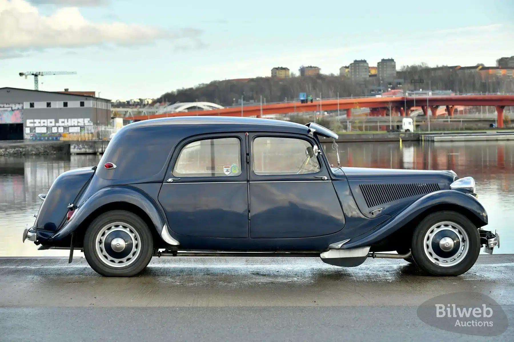 Seitenansicht eines Citroën B11 Traction Avant von 1956 mit dunklem Blau, aufgenommen am Wasser vor urbanem Hintergrund.
