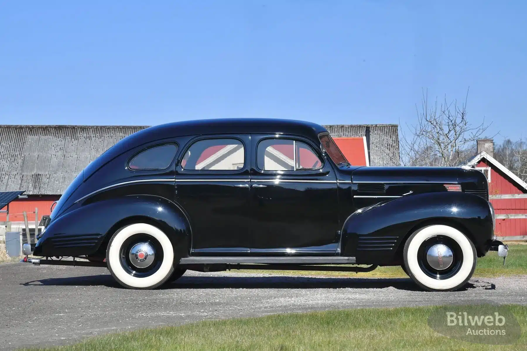 Seitenansicht eines schwarzen Dodge Senior Touring Sedan von 1939 mit weißwandreifen auf einem ländlichen Hof vor Scheunen.