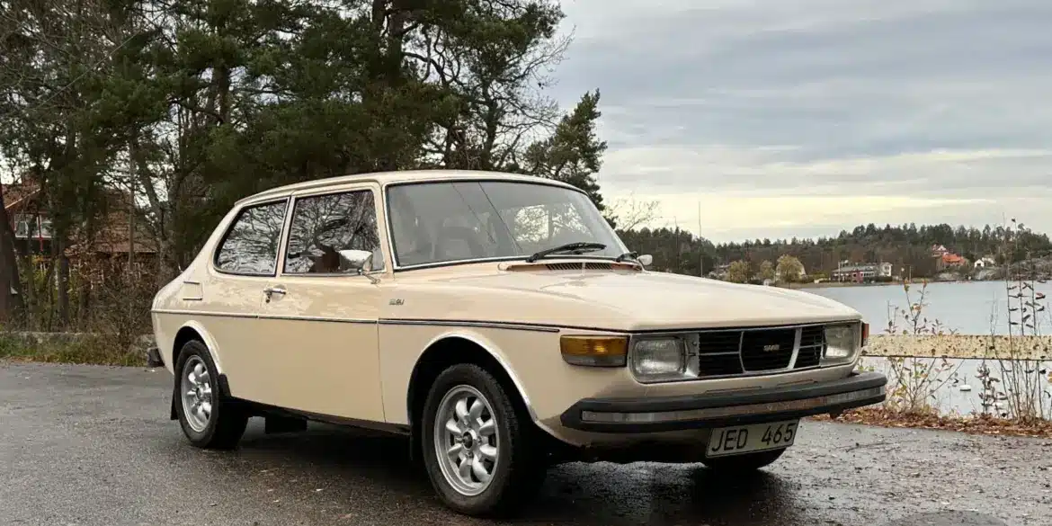 Saab 99 GL, Baujahr 1976, in beige auf einem Parkplatz vor herbstlicher Kulisse.