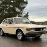 Saab 99 GL, Baujahr 1976, in beige auf einem Parkplatz vor herbstlicher Kulisse.