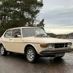 Saab 99 GL, Baujahr 1976, in beige auf einem Parkplatz vor herbstlicher Kulisse.
