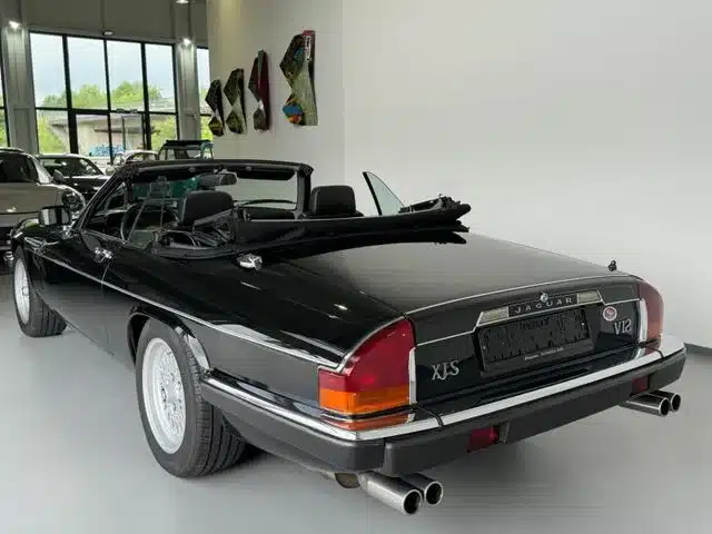 Heckansicht eines schwarzen Jaguar XJS V12 Cabriolets mit geöffnetem Verdeck, Arden-Alufelgen und Vierrohr-Abgasanlage in einer Ausstellungshalle.