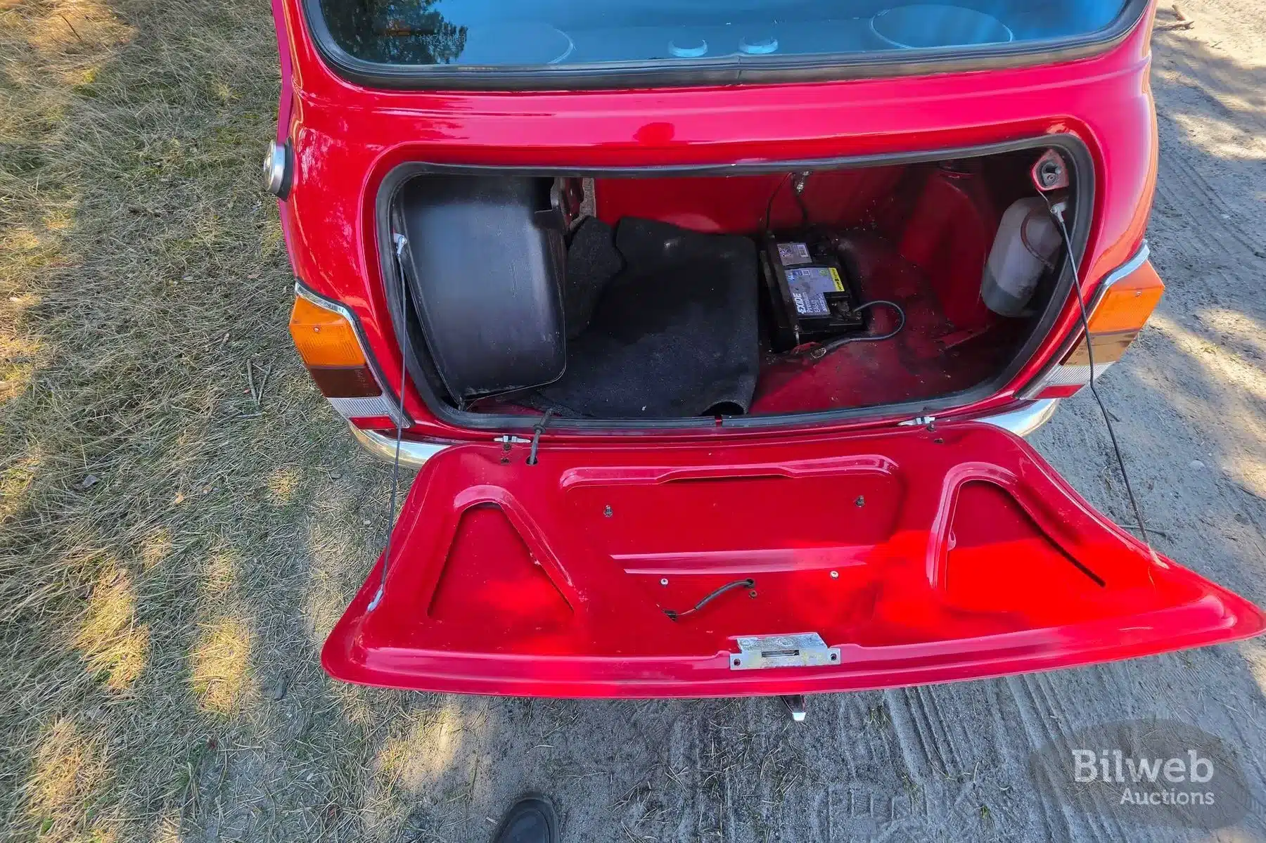 Blick in den geöffneten Kofferraum eines roten Rover Mini Cooper von 1997 mit sichtbarer Batterie und Ersatzrad.
