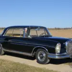 Schwarzes Mercedes-Benz 300 SE Coupé der Baureihe W112, Baujahr 1964, auf einem Feldweg vor offenem Landschaftshintergrund
