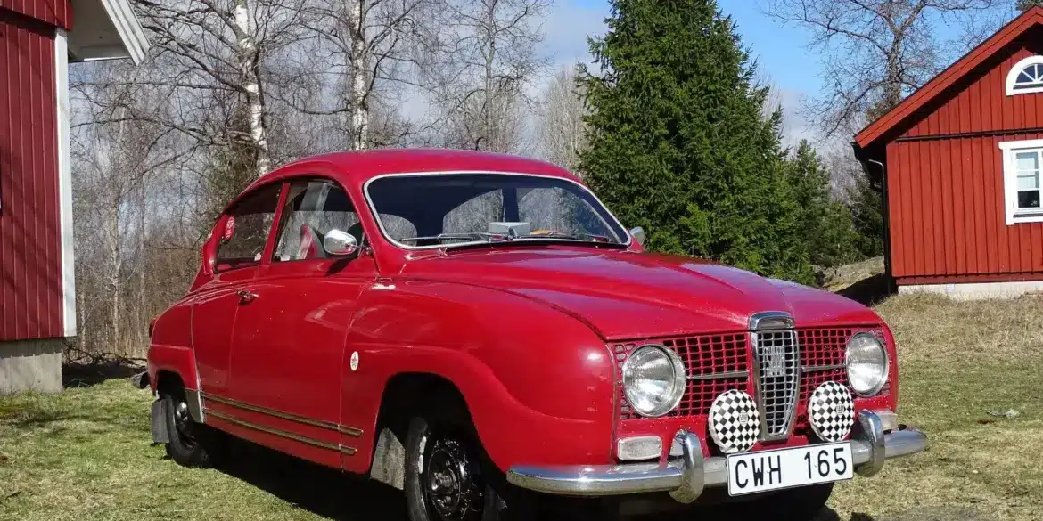 Roter Saab 96 Monte Carlo 850 Zweitakter von 1966 mit Patina, im Freien vor roten Holzhäusern fotografiert.