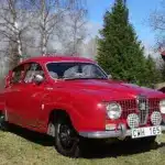Roter Saab 96 Monte Carlo 850 Zweitakter von 1966 mit Patina, im Freien vor roten Holzhäusern fotografiert.