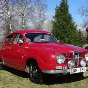 Roter Saab 96 Monte Carlo 850 Zweitakter von 1966 mit Patina, im Freien vor roten Holzhäusern fotografiert.