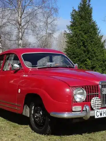 Roter Saab 96 Monte Carlo 850 Zweitakter von 1966 mit Patina, im Freien vor roten Holzhäusern fotografiert.
