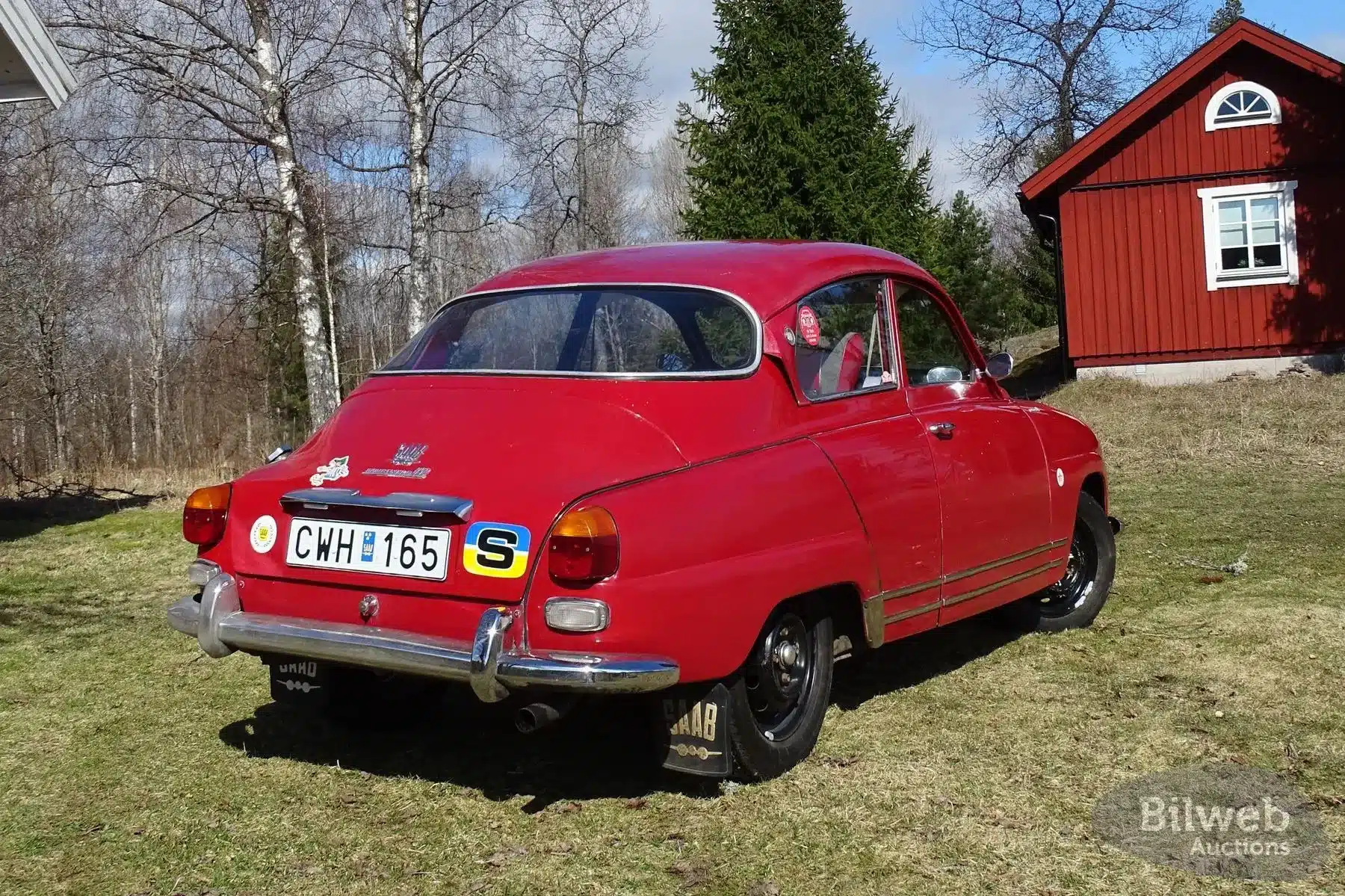Roter Saab 96 Monte Carlo 850 2-Takt von 1966 mit sichtbarer Patina, seitlich von hinten aufgenommen, vor einem roten Haus in ländlicher Umgebung