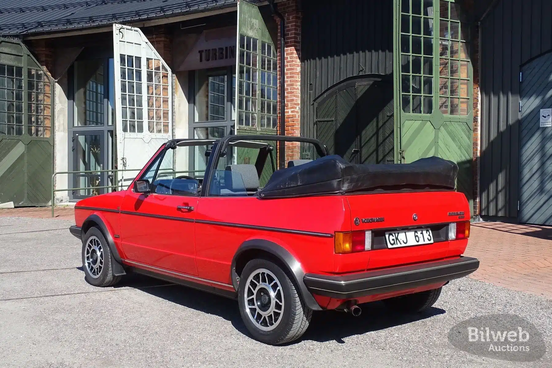 Seitenansicht eines roten VW Golf Cabriolet 1.8 GLI Baujahr 1985 mit geöffnetem Verdeck, im Hintergrund historische Industriegebäude