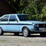 Hellblauer VW Passat TS 1500 von 1974 in Seitenansicht, mit Doppelscheinwerfern und Chromstoßstangen, aufgenommen vor Waldkulisse.
