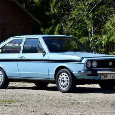 Hellblauer VW Passat TS 1500 von 1974 in Seitenansicht, mit Doppelscheinwerfern und Chromstoßstangen, aufgenommen vor Waldkulisse.