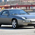 Originaler Porsche 928 S4 V8 von 1988 in silbergrauer Lackierung, seitlich vor urbaner Kulisse am Wasser abgestellt