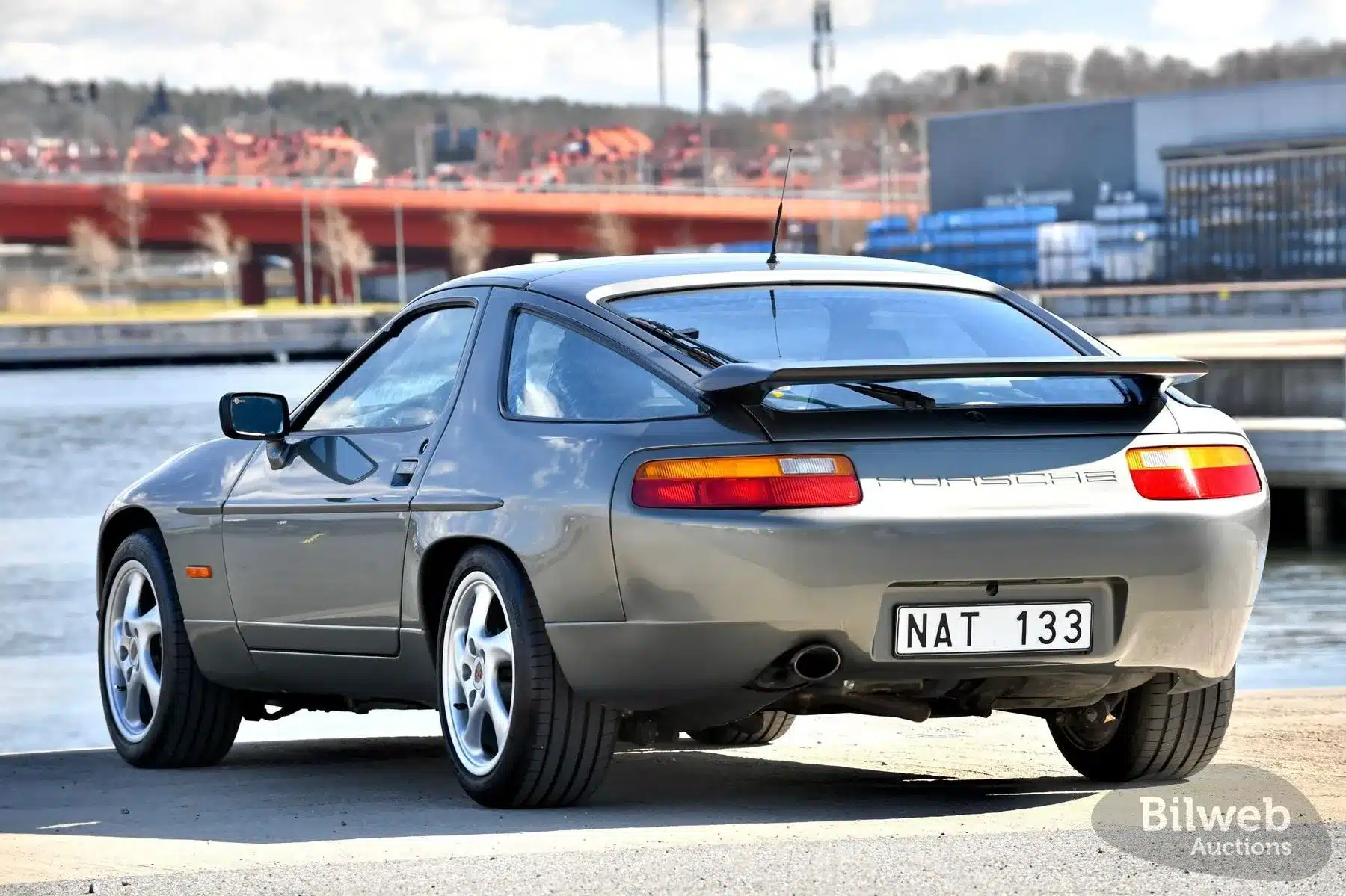 Porsche 928 S4 V8 von 1988 in Silber, Ansicht von hinten, im Freien an einem Wasserlauf, mit markanten Rückleuchten und Heckspoiler.