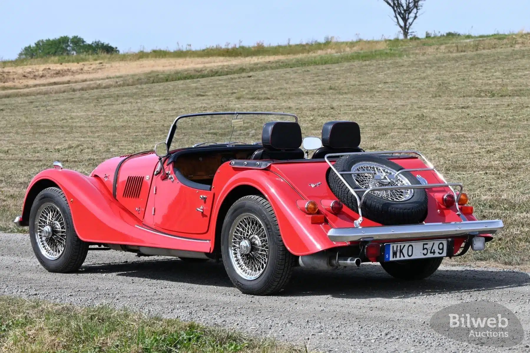 Roter Morgan 4/4 Roadster von 1998 mit Speichenrädern und Heckgepäckträger, geparkt auf einer Landstraße, aufgenommen von schräg hinten.