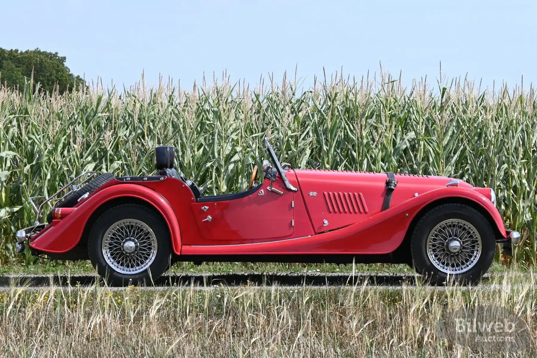 Seitenansicht eines roten Morgan 4/4 Roadster von 1998 auf einer Landstraße vor einem Maisfeld.