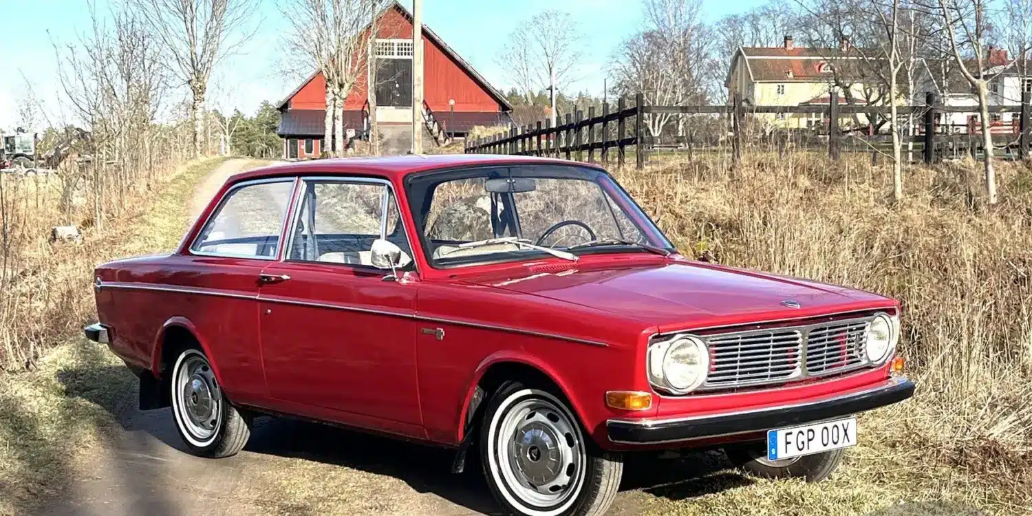 Volvo 142 B20 von 1969 in Rot, stehend auf einem ländlichen Weg vor Scheune.