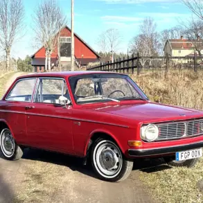 Volvo 142 B20 von 1969 in Rot, stehend auf einem ländlichen Weg vor Scheune.