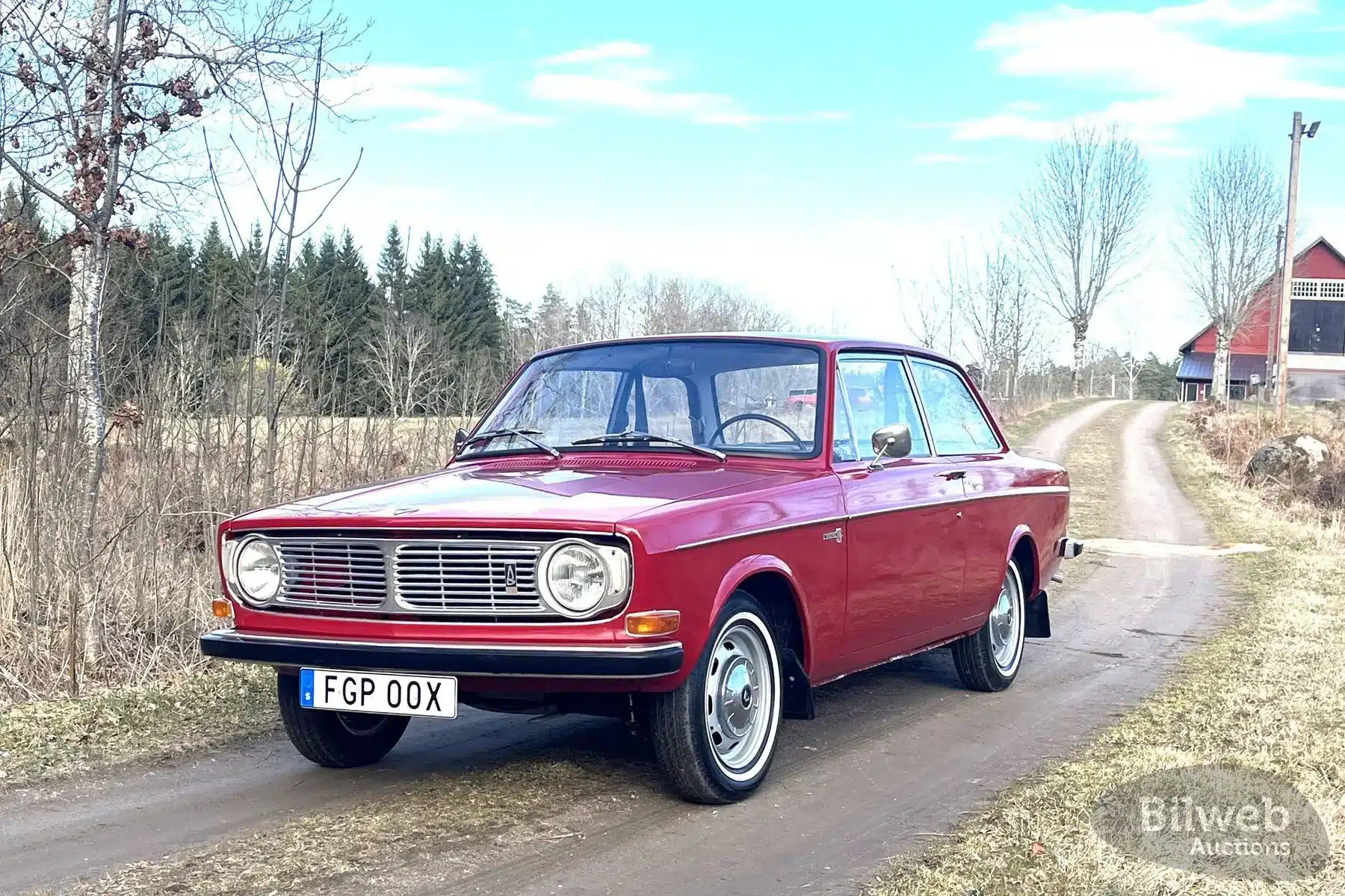 Roter Volvo 142 B20 von 1969 steht auf einer schmalen Landstraße, umgeben von Bäumen und Feldern.