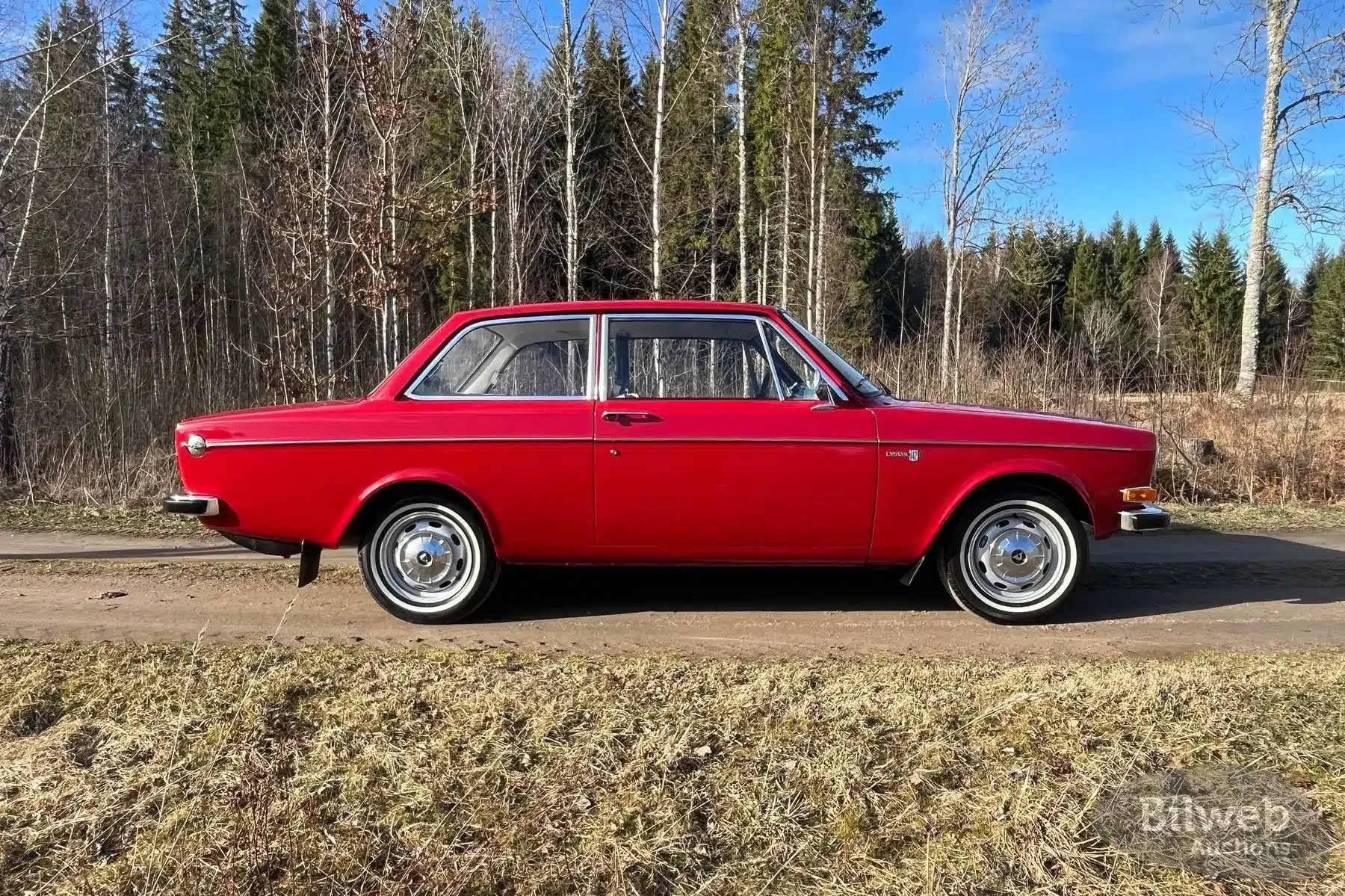 Roter Volvo 142 B20 von 1969, seitliche Aufnahme, stehendes Fahrzeug auf Landstraße mit Wald im Hintergrund.