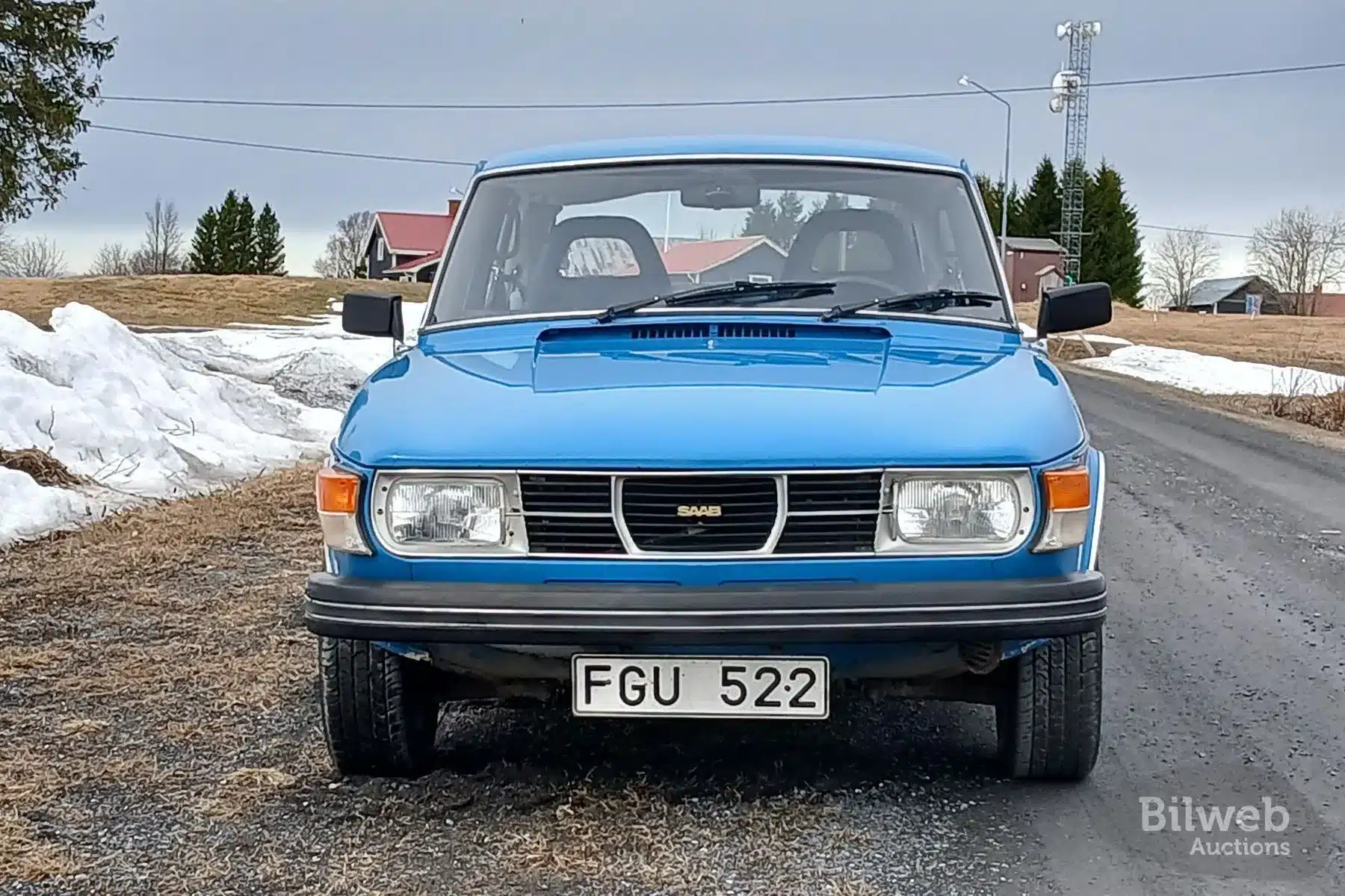 Frontansicht eines blauen Saab 99 GL 2.0 von 1977 mit schwedischem Kennzeichen auf einer Straße, umgeben von Schnee und ländlicher Landschaft.