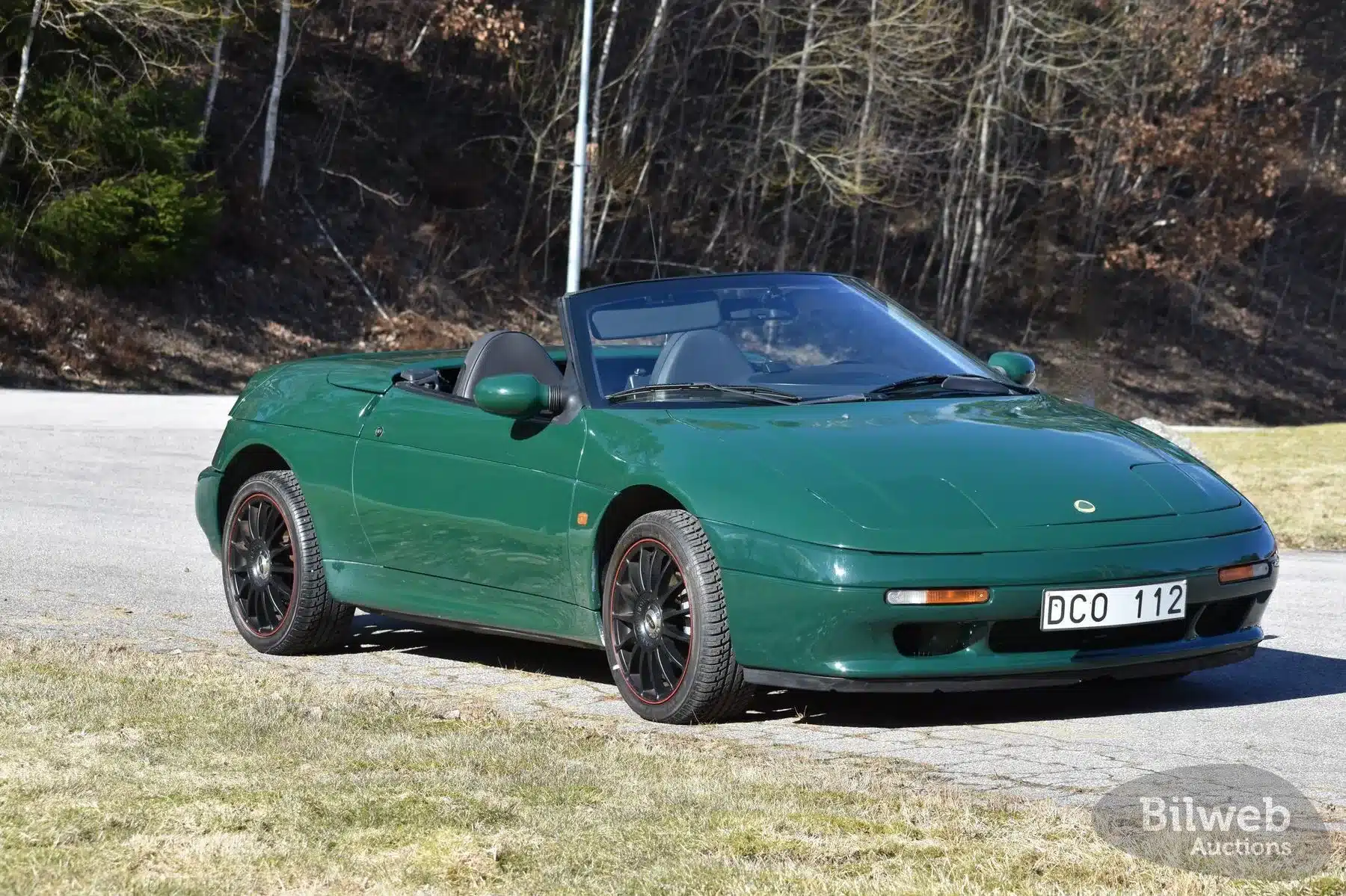 Dunkelgrüner Lotus Elan M100 1.6 Turbo 16V Cabrio von 1991 mit geöffnetem Verdeck, seitliche Frontansicht auf einer Straße