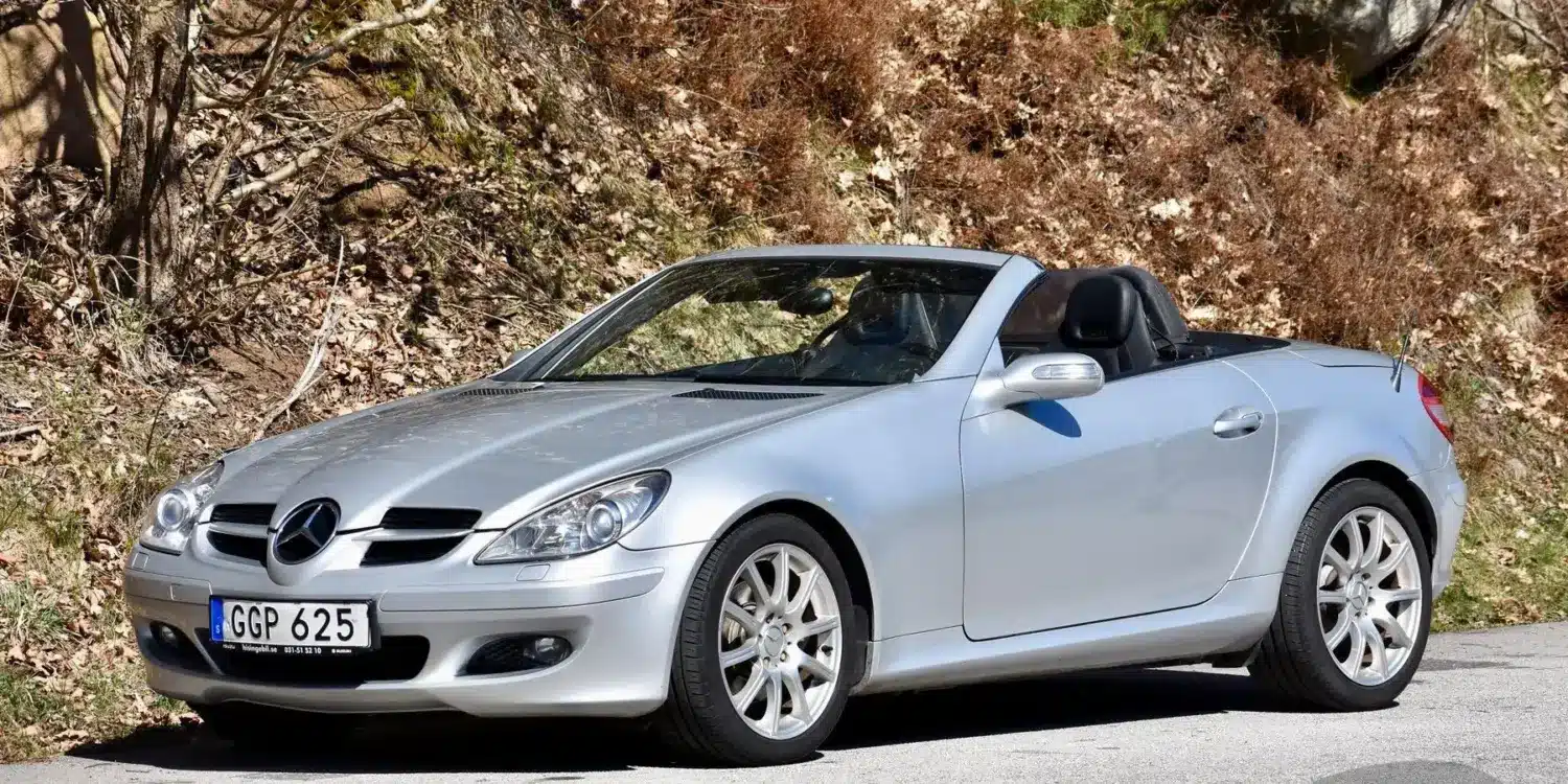 Silberner Mercedes-Benz SLK 280 (R171) mit geöffnetem Dach auf einer Straße vor herbstlicher Kulisse.