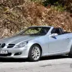 Silberner Mercedes-Benz SLK 280 (R171) mit geöffnetem Dach auf einer Straße vor herbstlicher Kulisse.