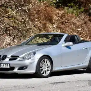 Silberner Mercedes-Benz SLK 280 (R171) mit geöffnetem Dach auf einer Straße vor herbstlicher Kulisse.