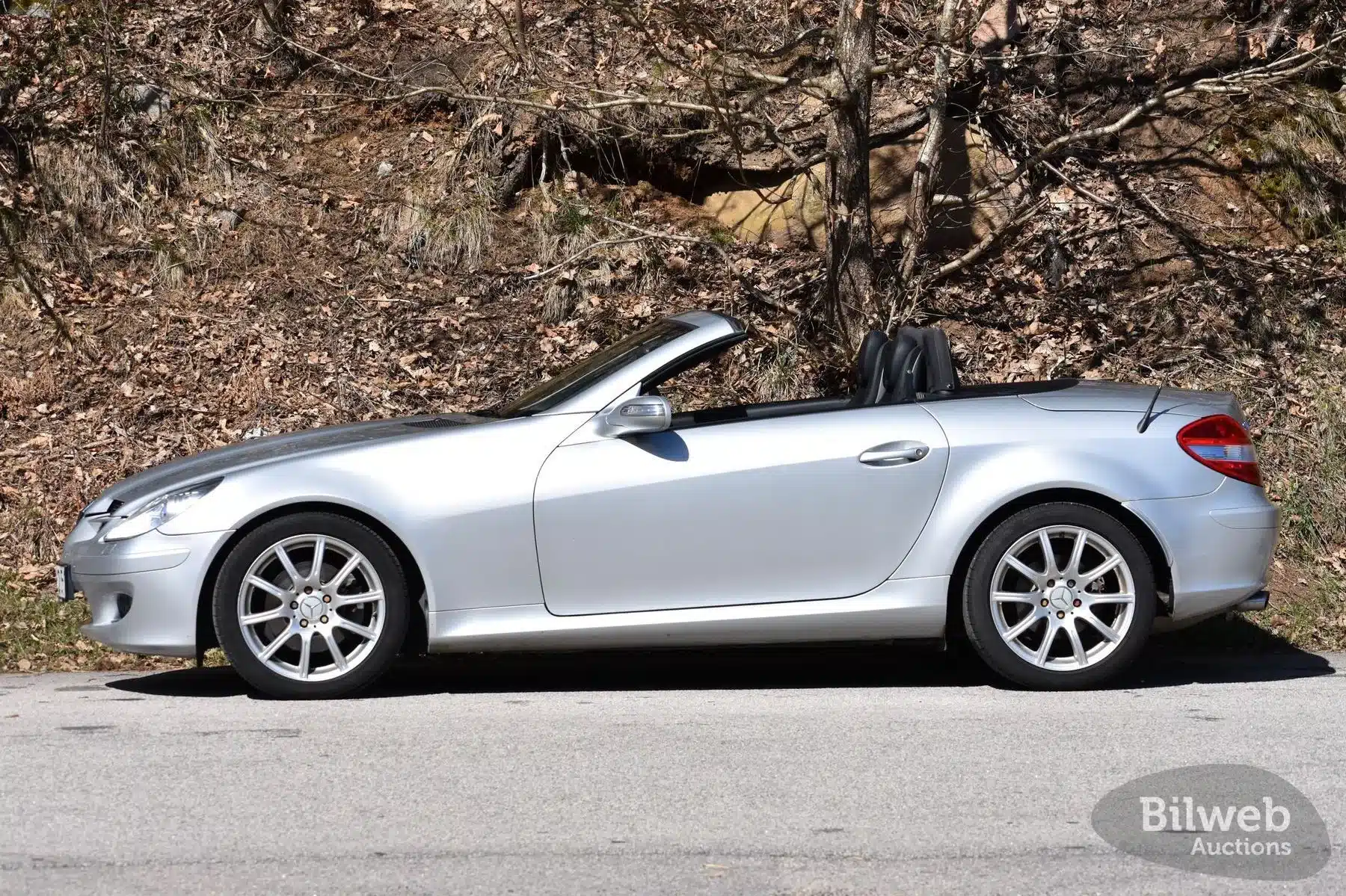 Silberner Mercedes-Benz SLK 280 R171 Roadster offen, seitlich auf einer Straße vor bewaldetem Hintergrund, Bilweb Auctions Logo