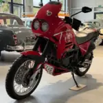 Triumph T900 Tiger in roter Lackierung, seitliche Ansicht in einer Halle mit klassischen Fahrzeugen im Hintergrund.