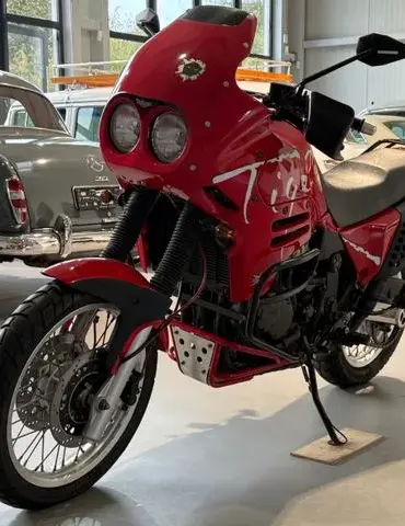 Triumph T900 Tiger in roter Lackierung, seitliche Ansicht in einer Halle mit klassischen Fahrzeugen im Hintergrund.