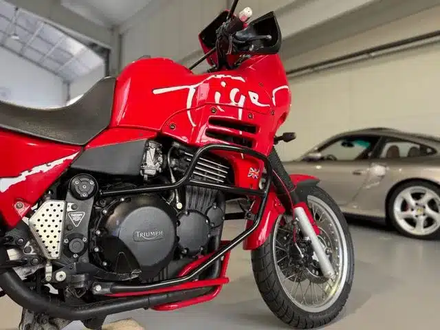 Rote Triumph T900 Tiger Reiseenduro, seitlich fotografiert, in einer Garage mit anderem Fahrzeug im Hintergrund.