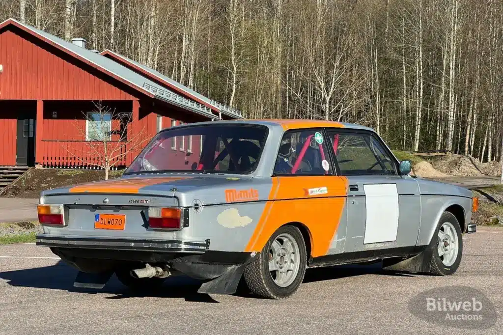 Volvo 142 Rallyeauto von 1970 mit orange-grauer Lackierung und sichtbaren Gebrauchsspuren, fotografiert auf einem Parkplatz vor einem roten Gebäude.
