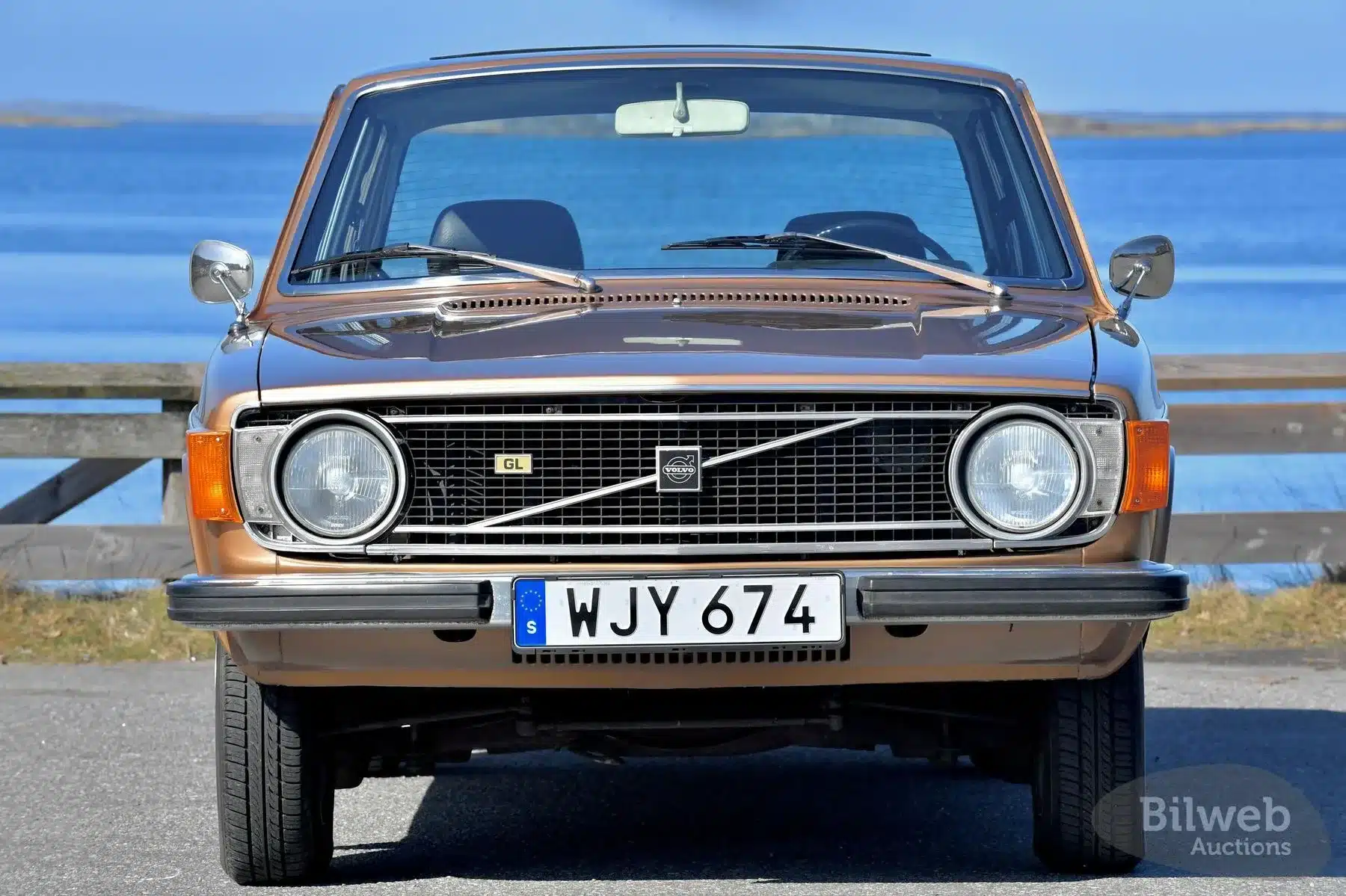 Frontal fotografierter brauner Volvo 144 GL B20 von 1973 mit schwedischem Kennzeichen vor Wasserlandschaft.