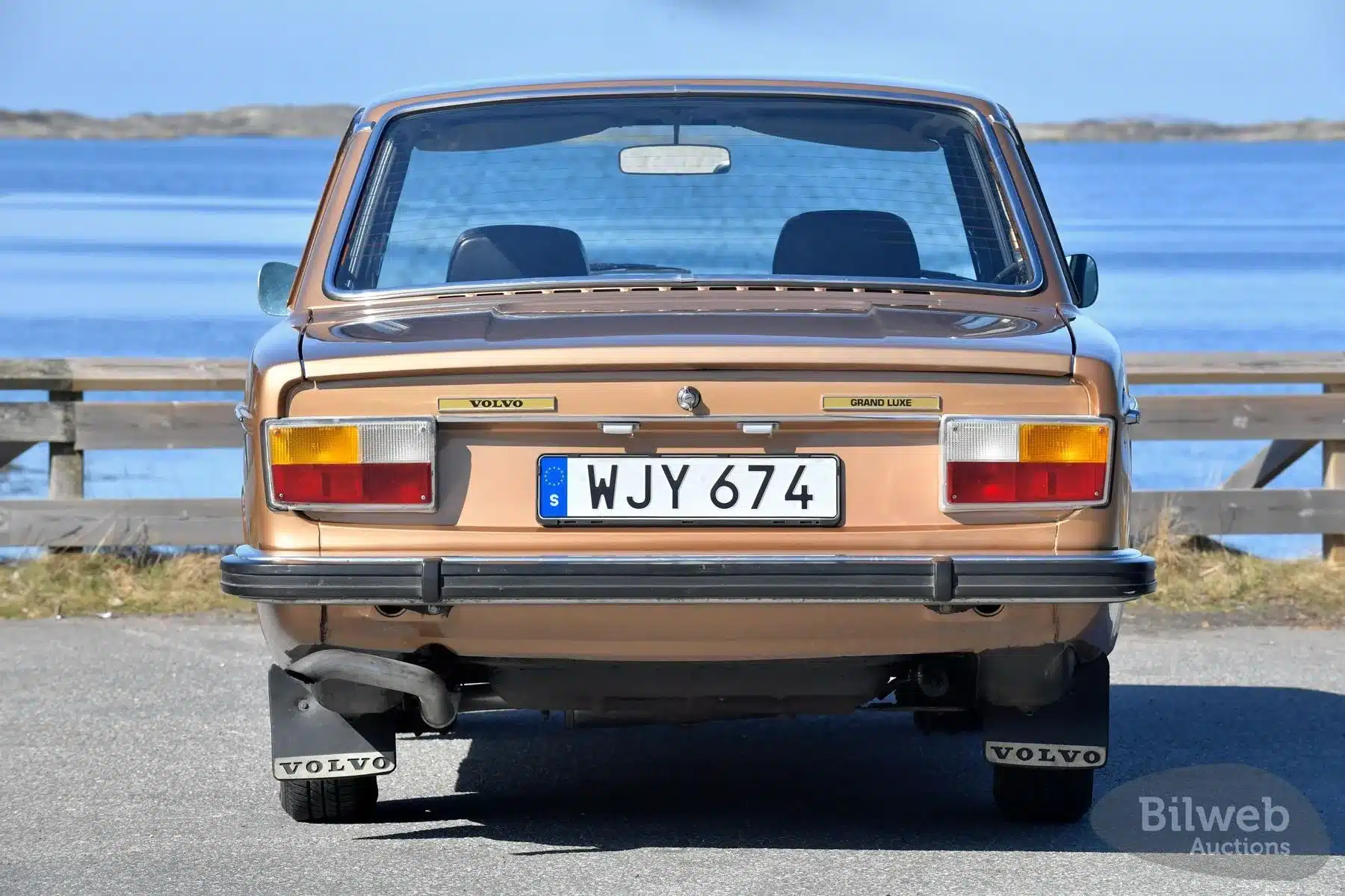 Rückansicht eines goldfarbenen Volvo 144 GL B20, Baujahr 1973, mit schwedischem Kennzeichen vor Gewässer.