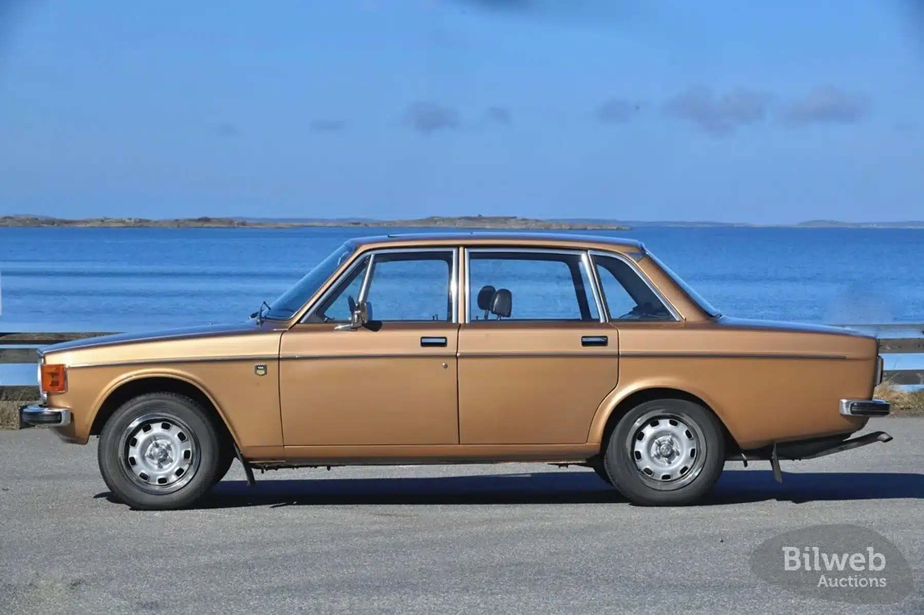 Seitenansicht eines Volvo 144 GL B20 von 1973 in Gold, geparkt vor einer Wasserlandschaft.