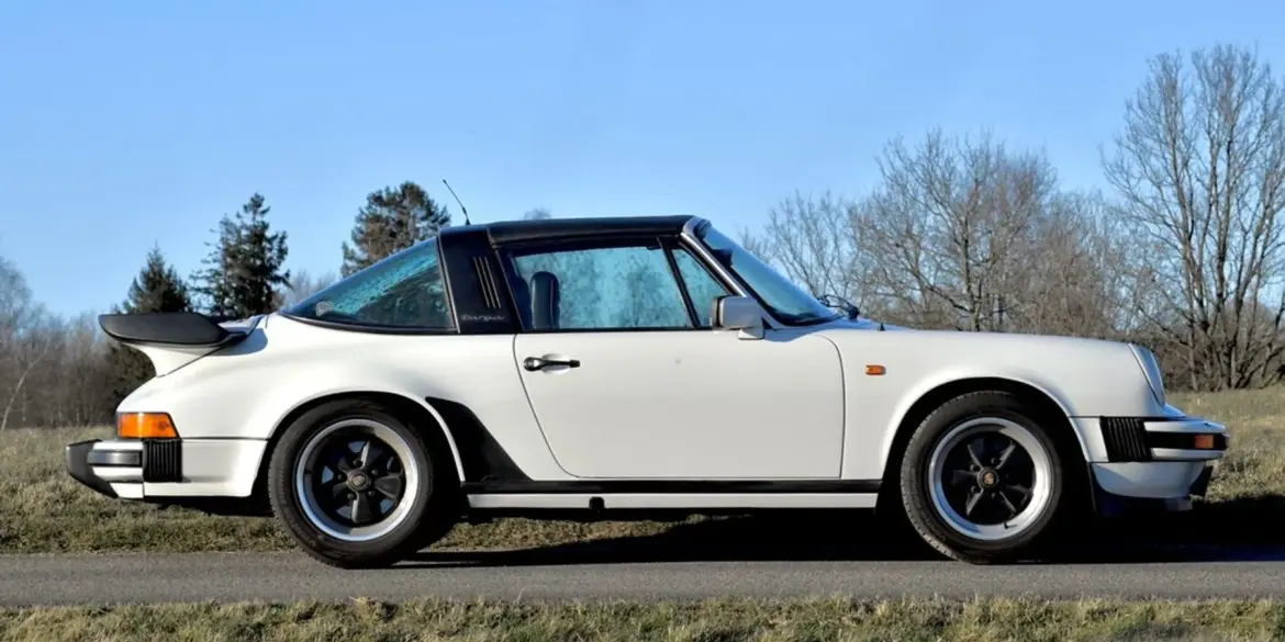 Weißer Porsche 911 SC Targa 3,0, Baujahr 1981, seitlich fotografiert, mit schwarzem Targa-Bügel und Fuchs-Felgen auf einer Landstraße vor natürlicher Kulisse.