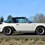 Weißer Porsche 911 SC Targa 3,0, Baujahr 1981, seitlich fotografiert, mit schwarzem Targa-Bügel und Fuchs-Felgen auf einer Landstraße vor natürlicher Kulisse.